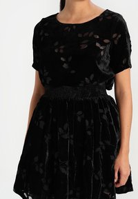 Robe en velours noir avec un motif floral, des manches courtes, une taille froncée, une jupe évasée et des détails transparents.