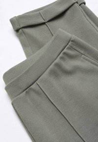 Gros plan sur un pantalon en tissu texturé vert olive plié, montrant la taille et les détails des coutures sur un fond blanc.
