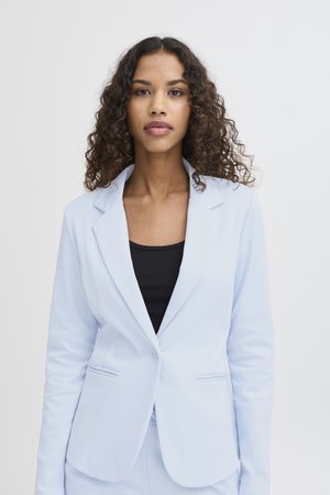 Junge Frau mit lockigem Haar trägt ein hellblaues Sakko über einem schwarzen Oberteil und steht vor einem schlichten weißen Hintergrund.