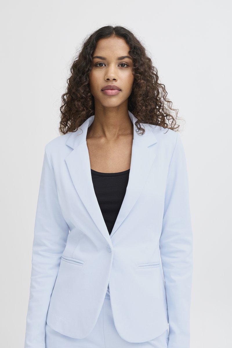 Mlada žena s kovrčavom kosom nosi svijetloplavi blejzer preko crne majice, stojeći ispred čistog bijelog zida.