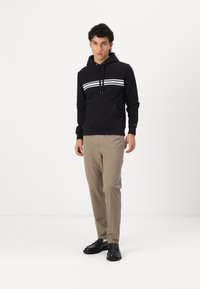 Sort hættepullover med hvide striber, kengurulomme og justerbar snor. Sammen med beige bukser og sorte sko.