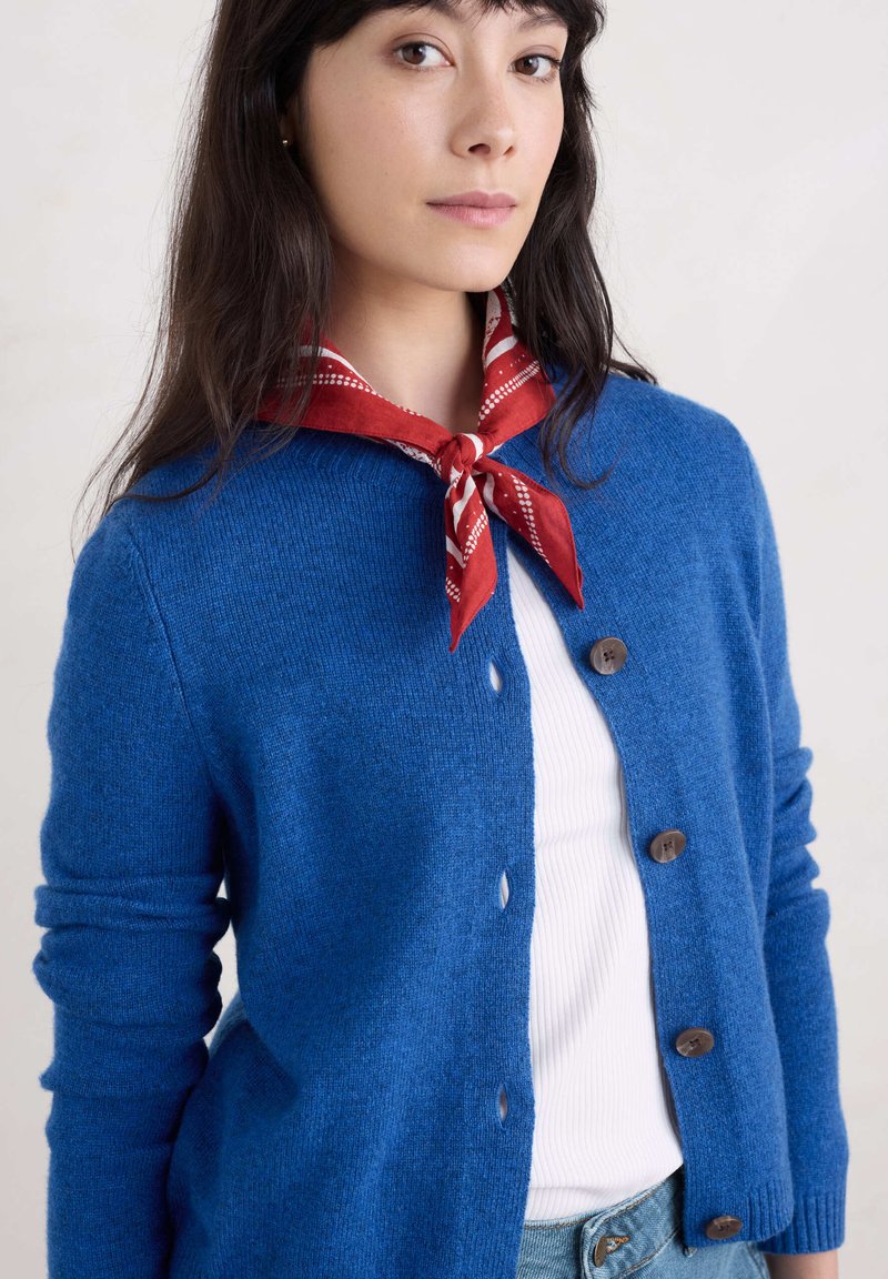 Blå kardigan i mjukt stickat material med rund halsringning, knappstängning och ribbade ärmslut. Bärs över en vit topp med en röd och vit mönstrad scarf.