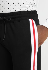 Persoon in zwart sweatpants met elastische tailleband en witte en rode zijstrepen, hand in zak, gecombineerd met een wit met stippen topje.