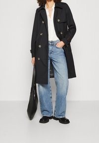 Svart trenchcoat med dubbelknäppt design, silverknappar och justerbara ärmar, i kombination med ljusblå vida jeans och en svart tote-väska.