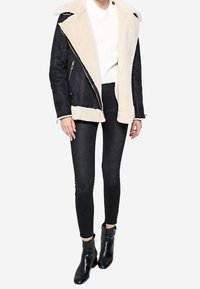Kvinde iført sort lammeskindjakke med cremefarvet foer, hvid sweater, sorte skinny jeans og sorte ankelstøvler, stående mod en hvid baggrund.