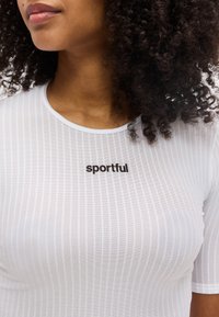 Κοντινό πλάνο ενός ατόμου με σγουρά μαλλιά που φοράει άσπρο ριπ μπλουζάκι με την λέξη "sportful" τυπωμένη μαύρα στο στήθος.
