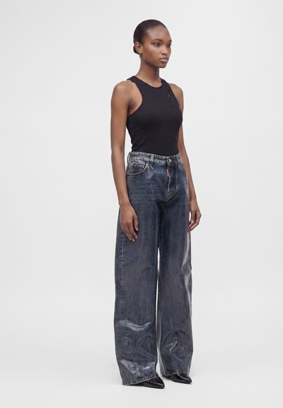 Jeune femme debout portant un haut noir sans manches, un jean bleu délavé à jambes larges et des chaussures noires pointues sur un fond clair uni.