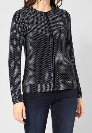 Kvinde iført en sort og hvid mønstret langærmet lynlåsjakke med mørkeblå jeans mod en ensfarvet lys baggrund.