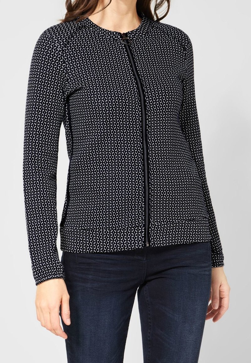 Femme portant une veste zippée à manches longues à motif noir et blanc avec un jean bleu foncé sur un fond clair uni.