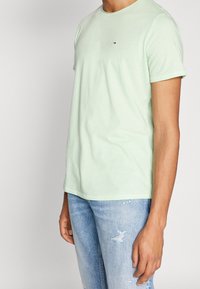 Světlezelené bavlněné tričko s krátkými rukávy, kulatým výstřihem a malým logem na hrudi, v kombinaci s opranými modrými denimovými džíny.