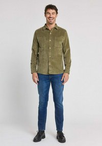 Chemise en velours côtelé vert olive avec deux poches avant et fermeture par boutons, associée à un jean bleu et des chaussures noires. Dotée d'un col et de manches longues.
