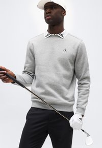 Άνδρας που φοράει γκρι φούτερ CK, λευκό γάντι του γκολφ και λευκό καπέλο, κρατώντας ένα μπαστούνι του γκολφ μπροστά σε απλό φόντο.