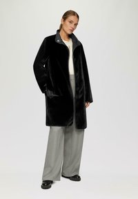 Cappotto lungo nero in peluche con colletto grigio, dotato di chiusura a zip e due tasche laterali, indossato sopra pantaloni larghi grigi chiari.