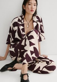 Femeie așezată pe podea, purtând un costum larg cu modele bej și violet închis, balerini negri și un colier cu pandantiv.