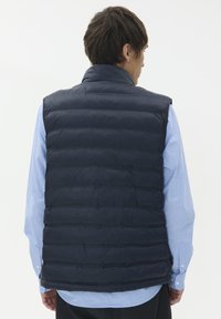 Marinblå pufferväst med vertikalt quiltat mönster, hög krage och lättviktsmaterial. Bärs över en ljusblå skjorta. Enkel och funktionell.