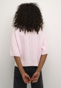 Różowy sweter na drutach z krótkimi rękawami, zdobiony wzorem liści, z ściągaczami na mankietach i luźnym krojem. Noszony z ciemnymi dżinsami, widziany od tyłu.