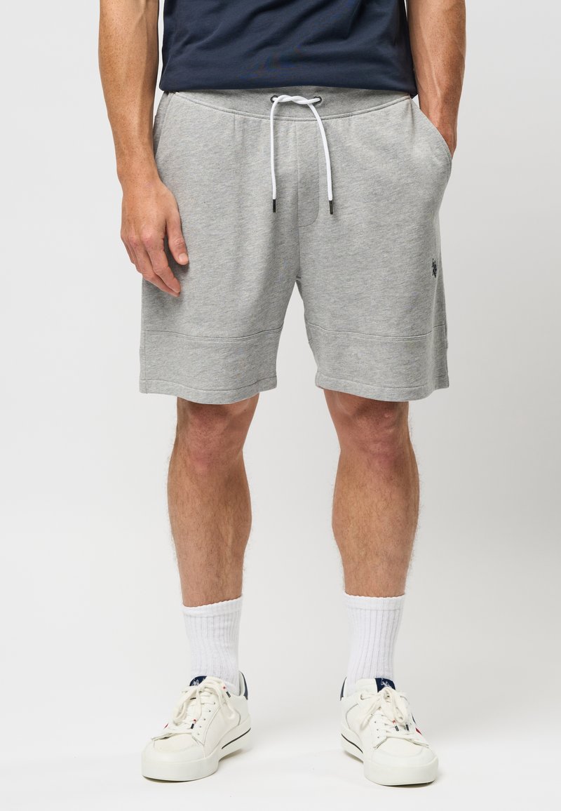 Shorts en coton gris avec taille élastique et cordon de serrage, poches latérales et un petit logo sur le côté. Associés à des baskets blanches et des chaussettes.