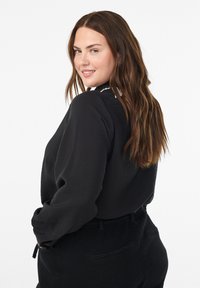 Donna con lunghi capelli castani, che indossa una maglietta nera a maniche lunghe e pantaloni neri, che sorride e guarda oltre la spalla su uno sfondo bianco.
