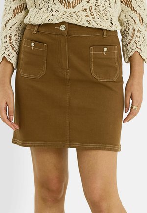 Minifalda marrón con botones delanteros y dos bolsillos parche con botón, combinada con un top de manga larga de crochet color crema y anillos en una mano.