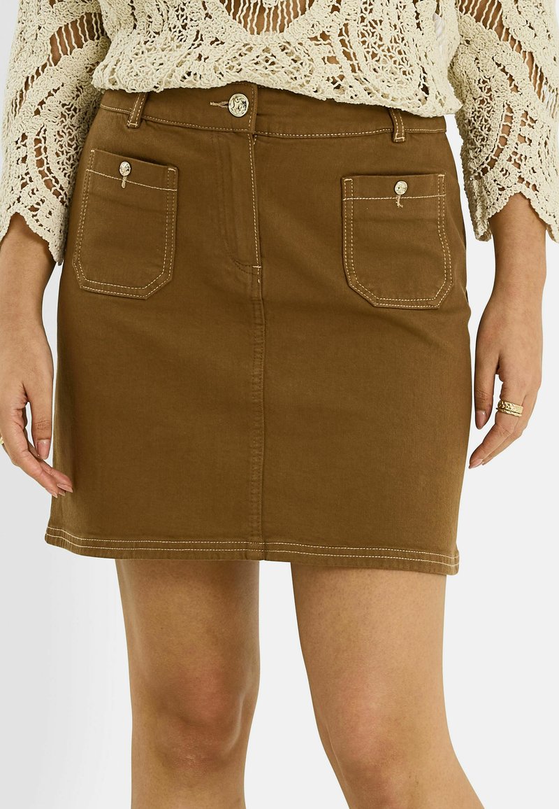 Minifalda marrón con botones delanteros y dos bolsillos parche con botón, combinada con un top de manga larga de crochet color crema y anillos en una mano.