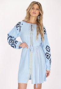 Vaaleansininen mekko, jossa on pitkät hihat ja mustaa geometristä brodeerausta. Suunnittelussa on nauhakuja ja vyötärölle solmittava vyö.