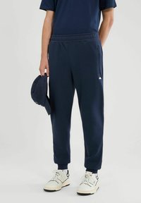 Osoba ubrana w granatowe spodnie dresowe, czarną koszulkę, białe trampki, trzyma pasującą granatową czapkę na jasnym, jednolitym tle.