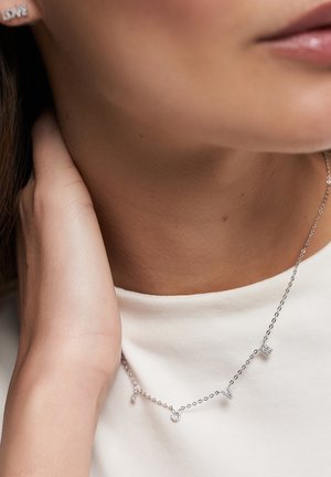 Silver chain necklace featuring small geometric pendants with varied textures and accents. Worn over a white top, resting along the neck.