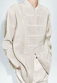Camicia button-up di un beige chiaro con colletto alla mandarina, caratterizzata da righe verticali e una vestibilità rilassata, realizzata in un tessuto testurizzato.
