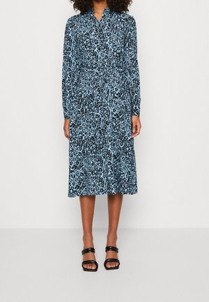 Robe midi bleue à fleurs avec un col boutonné, des manches longues et une taille élastiquée. Associée à des sandales noires à brides.