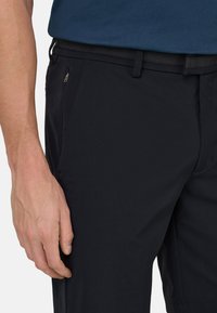 Mano maschile posata di lato, che indossa pantaloni neri con tasca con cerniera e camicia blu infilata, ritagliata a livello della vita e della coscia.