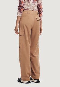 Pantaloni cargo beige con vita alta e tasche laterali. Il tessuto è liscio con una leggera lucentezza. Abbinati a calzature sportive nere.