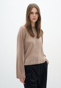 Maglione a maniche lunghe di colore marrone chiaro, realizzato in tessuto morbido, caratterizzato da una vestibilità rilassata e un design minimal; indossato con pantaloni neri a gamba larga.