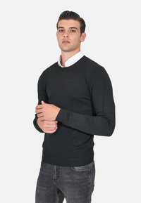 Maglione nero a maglia con scollatura rotonda e polsini a coste, abbinato a una camicia bianca con colletto sotto. Il modello posa con le mani unite.