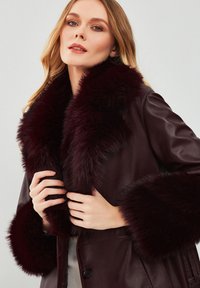 Giacca di pelle bordeaux con un morbido colletto e polsini in pelliccia. Presenta grandi bottoni e una texture liscia, che mette in mostra un design moderno.