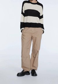 Maglione a righe neri e crema, taglio ampio. Pantaloni beige dalla forma rilassata, con tasche laterali. Scarpe nere robuste.