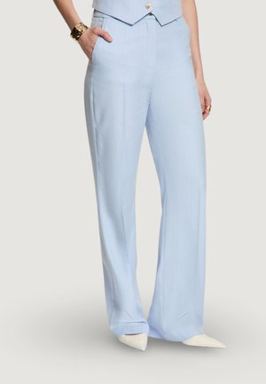 Mujer con pantalones de tiro alto y pierna ancha color azul claro con bolsillos laterales, combinados con un chaleco a juego y zapatos blancos de punta.