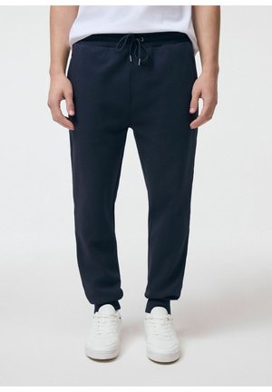 Mężczyzna w czarnych spodniach dresowych ze ściągaczem i białych trampkach, stojący na tle jednolitego jasnego tła.