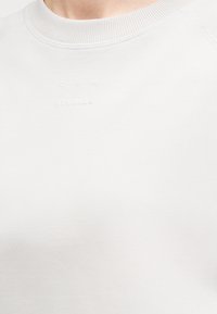 Gros plan sur un sweat-shirt blanc à col rond avec le texte en relief subtil "CPH STUDIOS" au niveau de la poitrine.