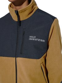 Person trägt eine braune und dunkelgraue Fleecejacke mit Frontreißverschluss und dem Schriftzug „HALO 196819721981“ auf der Brust.