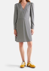 Abito a scacchi bianco e nero con maniche lunghe a palloncino e scollatura a V, abbinato a sneakers gialle con lacci bianchi e suole marroni.