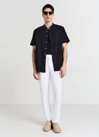 Chemise noire à manches courtes boutonnée portée sur un débardeur noir, associée à un pantalon blanc et des chaussures beige à enfiler. Lunettes de soleil en accessoire.