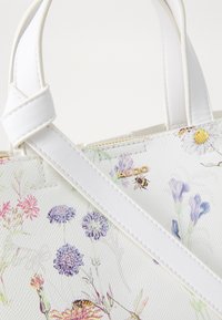 Sac à main avec imprimé floral blanc, fabriqué à partir d'un matériau texturé, avec un logo en métal doré et une sangle blanche. Le design comprend des fleurs colorées et des abeilles.
