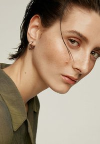 Aro de plata con un diseño texturizado, llevado en la oreja izquierda. El sujeto tiene el cabello corto y húmedo y viste una camisa verde oliva.
