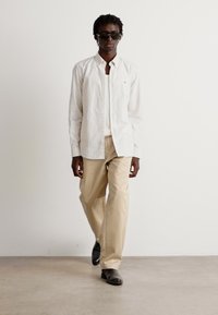 Camicia a righe bianche con bottoni, pantaloni beige e scarpe nere. Il modello indossa occhiali da sole, con una silhouette semplice e classica. Tessuto dalla texture liscia.