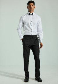 Cămașă albă cu nasturi negri, asociată cu pantaloni negri și un papion negru; pantofi negri lustruiți completează look-ul formal.