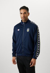 Veste de survêtement zippée marine en tissu léger. Comprend un logo, trois étoiles et des accents rayés le long des manches. Poignets et ourlet élastiques.