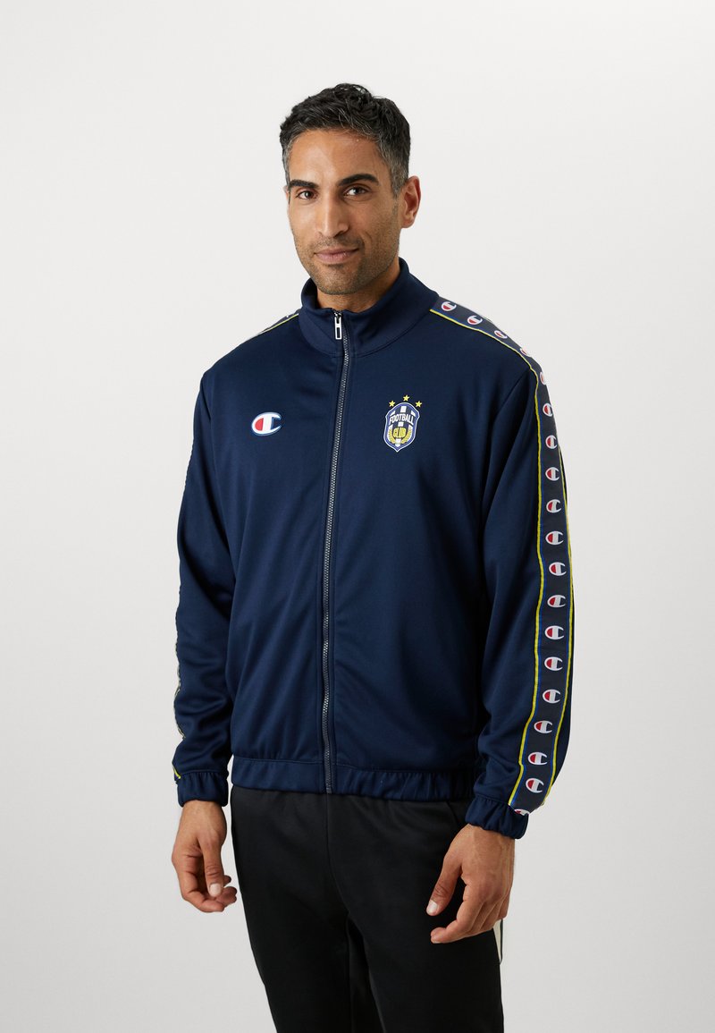 Marineblå zip-up træningsjakke lavet af letvægtigt stof. Har logo, tre stjerner og stribede detaljer ned ad ærmerne. Elastiske cuffs og bund.