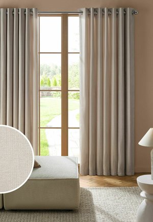 Des rideaux beige pendent sur une porte vitrée encadrée en bois avec vue sur un jardin, à côté d’un ottoman beige et d’une lampe de table sur un tapis texturé.