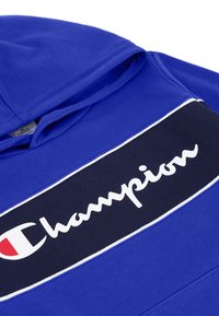Sudadera azul hecha de un tejido suave, con una gran banda negra que lleva el logotipo blanco "Champion", acentuada con ribetes blancos a lo largo de los bordes.