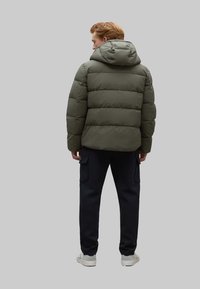 Chaqueta tipo puffer en verde oliva con capucha, con un diseño acolchado y una silueta compacta. Combinada con pantalones oscuros y zapatillas blancas.